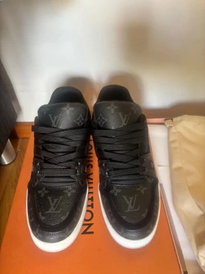 Louis Vuitton svarta monogram sneakers - Snygga svarta Louis Vuitton sneakers med klassiskt monogrammönster över hela skon. Skorna har vita sulor, svarta skosnören och är tillverkade i läder med detaljer i canvas. Perfekta för dig som vill ha en exklusiv och trendig look.