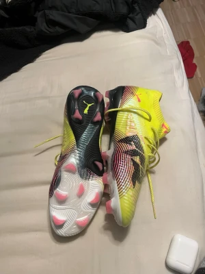 Puma gula fotbollsskor med dobbar neymar  - Säljer ett par grymma fotbollsskor från Puma i neon-gult med svarta och rosa detaljer. Skorna har snörning, högre strumpkrage och mönstrad ovansida för bättre bollkontroll. Dobbarna är rosa och vita, perfekta för spel på gräsplan. Riktigt snygg design som sticker ut på planen.