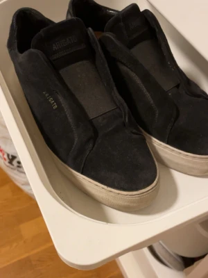 Axel Arigato - Säljer ett par svarta sneakers från Axel Arigato i mocka med minimalistisk design och vit sula. Diskret logga i guld på sidan och elastisk detalj över vristen för enkel påtagning. Perfekta för dig som gillar stilrena och cleana skor.