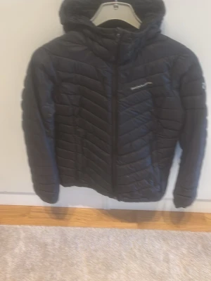 Svart dunjacka med huva från Peak Performance - Snygg svart dunjacka från Peak Performance med huva och dragkedja framtill. Jackan har ett quiltat mönster och diskret logga på bröstet. Perfekt för kalla vinterdagar och riktigt skön att bära. Klassisk passform och lätt att matcha.