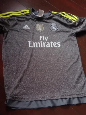Real Madrid Ronaldo matchtröja - Snygg grå Real Madrid matchtröja från Adidas med gula detaljer på axlarna och tryck av Ronaldo 7 på ryggen. Tröjan har klubbmärke, Adidas-logga och Fly Emirates-tryck på bröstet. Tillverkad i lätt och ventilerande material, perfekt för fotbollsträning.