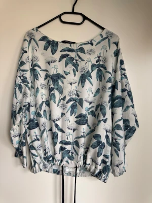 Blommig långärmad blus med dragsko - Snygg blus med vitt botten och grönt blommönster. Långärmad modell med lös passform och dragsko i nederkanten för en cool siluett. Tillverkad i ett lätt och mjukt tyg som känns skönt mot huden. Perfekt för dig som gillar en avslappnad men trendig stil.