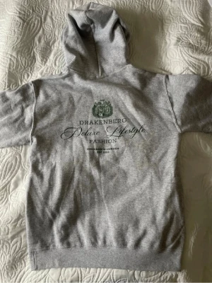 Grå hoodie från Drakenberg - Snygg grå hoodie från Drakenberg med tryck på ryggen och liten logga på bröstet. Klassisk modell med huva och känguruficka framtill. Tillverkad i mjukt bomullsmaterial som är skönt att ha på sig. Perfekt för en avslappnad och stilren look.