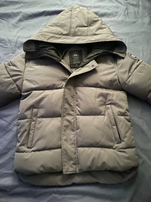 Grå dunjacka från Canada Goose - Snygg grå dunjacka från Canada Goose med huva och klassisk patch på ärmen. Jackan har raka fickor framtill och quiltad design som ger en modern look. Perfekt för kalla dagar tack vare det tjocka dunfodret och vindtäta materialet.