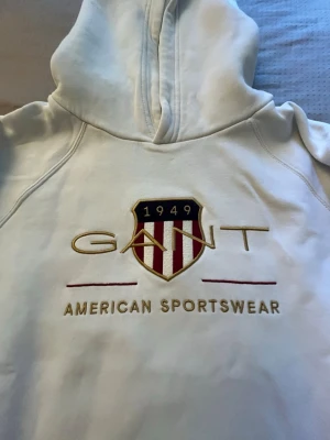 Vit hoodie från GANT  - Snygg vit hoodie från GANT med broderad logga och sköld på bröstet samt texten 'American Sportswear'. Hoodien har huva och är tillverkad i mjukt material, perfekt för en avslappnad och sportig stil.