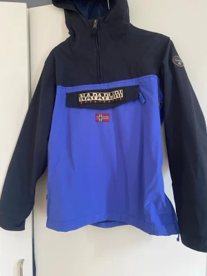 Napapijri vindjacka - Snygg blå och svart vindjacka från Napapijri med halv dragkedja, stor ficka framtill och norsk flagga som detalj. Jackan har huva och logga på ärmen. Perfekt för blåsiga dagar och streetwear-stil. 14y/162. Kontakta vid frågor!