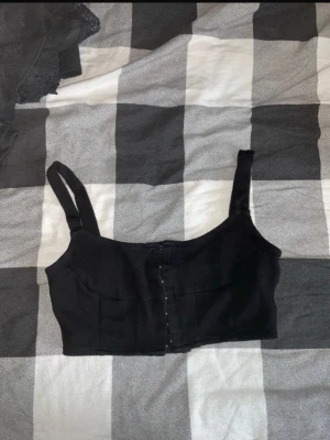 Svart croppad linnetopp - Snygg svart croppad linnetopp med breda axelband och knäppning framtill. Enkel och stilren design som passar perfekt till både jeans och kjol. Perfekt för dig som gillar minimalistisk stil.