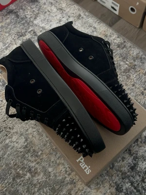 Christian Louboutin svarta spikes sneakers - Svarta sneakers i mocka från Christian Louboutin med ikoniska röda sulor och svarta nitar över tån och sidorna. Skorna har svarta snören och en clean low-top siluett som sticker ut med sin edgy design. Perfekta för dig som vill ha något unikt och vågat.