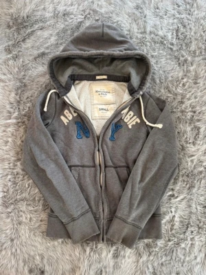 Grå hoodie från Abercrombie & Fitch - Säljer en grå zip-hoodie från Abercrombie & Fitch med broderade bokstäver i vitt och blått på bröstet. Hoodien har dragsko i huvan, mjukt foder och två fickor framtill. Perfekt för en avslappnad och cool stil. Kontakta vid ytligare frågor. Priset går att diskutera vid snabb affär. 