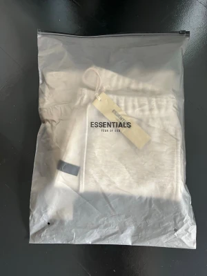 Grå Essentials Fear of God shorts - Snygga ljusgrå shorts från Essentials Fear of God med elastisk midja och långa snören framtill. Shortsen har fickor på sidorna och logga tryckt på vänster ben. Perfekta för en avslappnad och trendig stil.