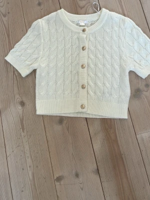 Vit kabelstickad kofta Gina Tricot - Kort, kabelstickad kofta i vit färg från Gina Tricot. Koftan har korta ärmar, rund halsringning och dekorativa guldfärgade knappar framtill. Ribbstickade muddar vid ärmslut och nederkant ger en snygg finish. Perfekt att styla över en topp eller klänning.