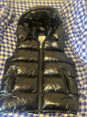 Svart g dunväst från Moncler - Säljer en svart, dunväst från Moncler. Västen har ett klassiskt Moncler-märke på bröstet och är tillverkad i ett quiltat material med dunfyllning. Perfekt för lager-på-lager och streetstyle.