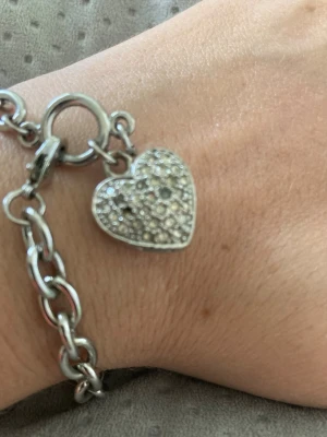 Silverfärgat armband med hjärthänge - Stilrent armband i silverfärgad metall med ett gnistrande hjärthänge dekorerat med små pärlor. Länkarna är kraftiga och armbandet har en stor ringlåsdetalj. Perfekt accessoar för att lyfta din look. Några diamanter har kommit av från hjärtat. 