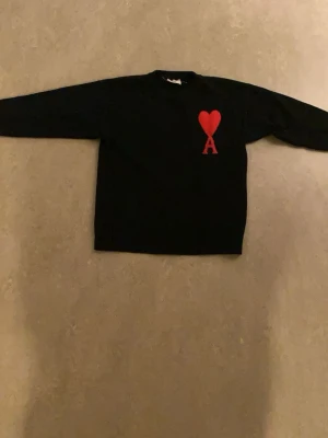 Svart stickad tröja Ami Paris - Säljer en svart stickad tröja från Ami Paris med röd hjärtlogga och bokstaven A på bröstet. Tröjan har rund halsringning och långa ärmar. Klassisk och stilren design som passar perfekt till många outfits.