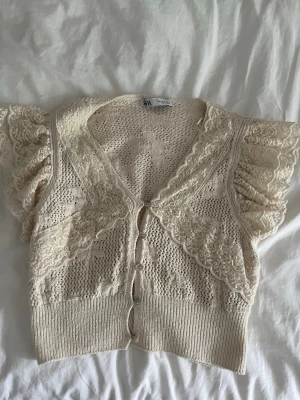 Krämvit spetskofta från Zara - Söt krämvit kofta från Zara med korta puffärmar och dekorativa spetsdetaljer. Koftan har v-ringning, knappar framtill och bred ribbad kant nedtill. Materialet är virkat och spetsen ger en romantisk känsla. Perfekt för att lyfta en enkel outfit.