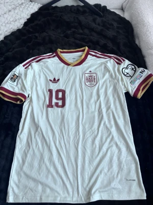 Vit Spanien VM 2026 bortaställ Adidas Lamine Yamal #19 - Säljer en vit Spanien fotbollströja från Adidas med vinröda och guldiga detaljer. Tröjan har nummer 19, broderat landslagsemblem och Adidas-logga på bröstet. Korta ärmar med matchmärken på båda ärmarna och Climalite-material för bra ventilation.