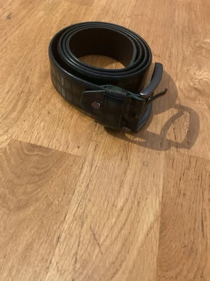 Mörk grönt bälte i skinn med krokoprägling - Snyggt mörkgrönt bälte i skinn med krokodilpräglat mönster och klassiskt metallspänne. Perfekt accessoar för att lyfta din outfit med en stilren och trendig detalj.