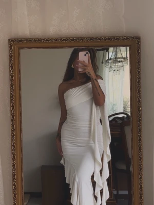 Vit oneshoulder långklänning - Säljer en elegant vit långklänning med oneshoulder-design och volangdetaljer längs sidan. Klänningen har en figurnära passform och är tillverkad i ett mjukt, stretchigt material som ger en snygg siluett. Köpt på shein, storlek S. Klänningen är enbart testad
