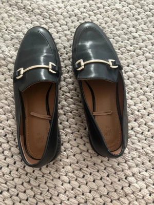 Svarta loafers med gulddetalj H&M - Stilrena svarta loafers från H&M i glansigt konstläder med guldfärgad metallspänne över foten. Klassisk design med rundad tå och låg klack. Perfekta för en trendig och enkel look.