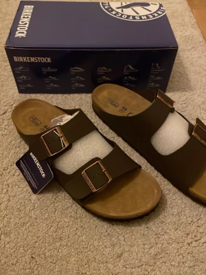 Bruna Birkenstock Arizona sandaler - Säljer ett par klassiska Birkenstock Arizona sandaler i mörkbrunt med två breda remmar och justerbara metallspännen i kopparfärg. Fotbädden är i mocka och sulan har korkdetaljer för extra komfort. Perfekta för en avslappnad och trendig stil.