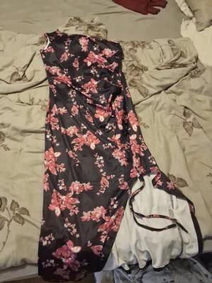 Svart blommig långklänning - Snygg svart långklänning med rosa och röda blommor. Klänningen har ärmlös design och ett mjukt, glansigt material som ger ett elegant intryck. Perfekt för dig som vill sticka ut med ett färgstarkt mönster och en feminin siluett.