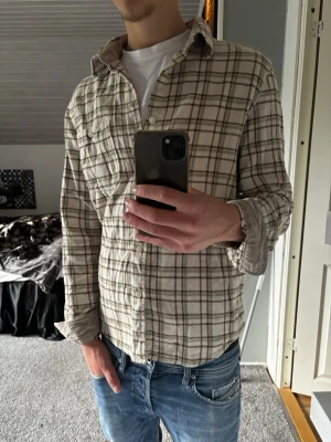 Beige rutig skjorta - Säljer en beige och brunrutig skjorta med klassisk krage och knappar framtill. Skjortan är tillverkad i mjuk bomull och har lång ärm. Perfekt för en avslappnad och stilren look.