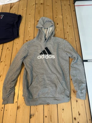 Grå hoodie från Adidas med logga - Snygg grå hoodie från Adidas med stor vit och svart logga på bröstet. Klassisk känguruficka framtill och mjuk insida. Tillverkad i bomullsmix och har en skön huva. Perfekt för chill eller träning.