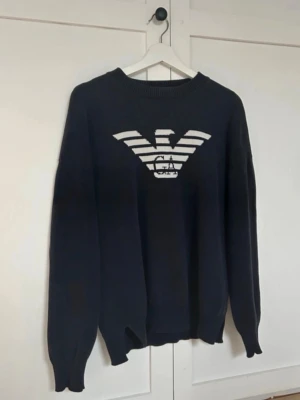 Svart stickad tröja Emporio Armani - Snygg svart stickad tröja från Emporio Armani med det ikoniska örnloggan i vitt på bröstet. Tröjan har rund halsringning, långa ärmar och ribbade muddar. Perfekt för dig som gillar stilrena och exklusiva plagg. Passar inte priset så är du välkommen att skicka ett pris förslag! (Nypris 2200)