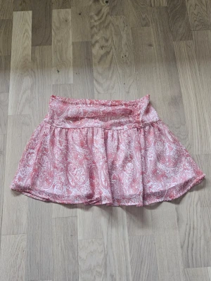 Gina Tricot rosa mönstrad sommarkjol - Jag säljer en söt, rosa och kort sommarkjol med vitt paisleymönster från Gina Tricot i storlek 146/152. Kjolen är knappt använd och därför i nyskick. 