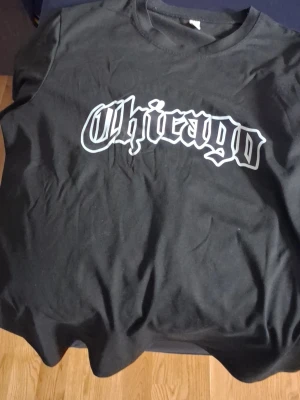 Svart t-shirt med Chicago-tryck - Snygg svart t-shirt med stort vitt Chicago-tryck i gotisk stil på bröstet. Klassisk rund halsringning och korta ärmar. Perfekt för dig som gillar streetwear och vill ha en enkel men stilren look.