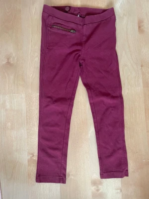Vinröda leggings med dragkedjedetalj storlek 104 - Snygga vinröda leggings med smal passform och en dekorativ dragkedja framtill. Mjukt och stretchigt material som sitter skönt. Perfekta till en avslappnad look och lätta att matcha med olika toppar.