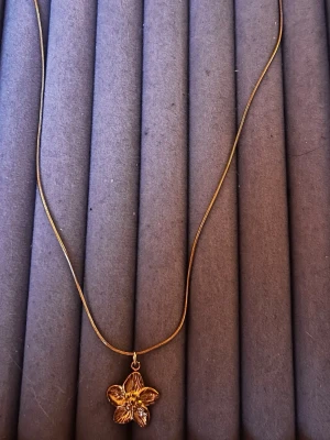 Guldigt halsband med blomma - Snyggt halsband i guldfärg med ett hängsmycke format som en blomma. Kedjan är tunn och smidig, och blomman har fina detaljer som ger ett elegant intryck. Perfekt för dig som gillar stilrena accessoarer med en touch av naturinspiration.