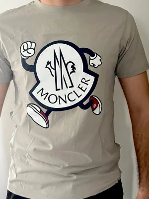 Beige Moncler t-shirt med stor logga - Snygg beige t-shirt från Moncler med en stor, tecknad logga framtill. T-shirten har korta ärmar och klassisk passform. Perfekt för dig som gillar streetstyle och vill sticka ut med ett coolt print. Helt ny, STORLEK L MEN PASSFORM FÖR MEDIUM OCKSÅ.