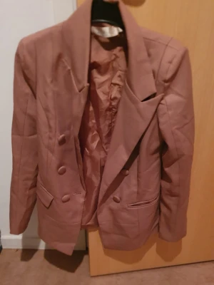 Kavaj - Stilren rosa kavaj med dubbelknäppning och breda slag. Kavajen har två fickor framtill och matchande tygklädda knappar. Perfekt för dig som vill ha en trendig och klassisk look. Det är i kinesiskt storlek men motsvarar svensk storlek 34 eller 36.