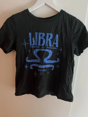 Svart t-shirt med blått Libra-tryck - Cool svart t-shirt med blått tryck av stjärntecknet Libra och texten 'Charismatic Peacemaker Artistic'. T-shirten har rund hals och korta ärmar, perfekt för dig som gillar astrologi och vill sticka ut lite extra.