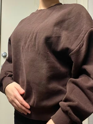 Brun oversized hoodie - Mörkbrun oversized hoodie med rund halsringning och ribbade muddar vid ärmslut och nederkant. Tillverkad i mjukt bomullsliknande material som känns skönt mot huden. 
