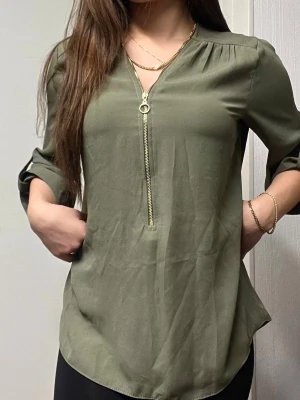 Olivgrön blus med dragkedja - Stilren olivgrön blus med lång ärm och guldfärgad dragkedja framtill. Blusen har en lätt rundad nederkant och mjukt, följsamt tyg som ger en avslappnad men ändå chic look. Perfekt att matcha med både jeans och kjol.