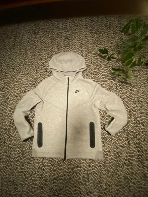 Ljusgrå Nike tech - Snygg ljusgrå hoodie från Nike med svart dragkedja och två svarta fickor framtill. Klassisk Nike-logga på bröstet och huva för extra stilpoäng. Tillverkad i mjukt material som känns skönt mot huden. Perfekt för dig som gillar sportig och clean look.
