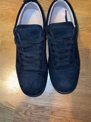 Svarta sneakers  - Säljer ett par svarta sneakers  med snygg broderad logga på hälen. Skorna har klassisk låg profil, svarta skosnören och ovandel i mocka. Yttersulan är svart med röd undersida. Perfekt för dig som gillar stilrena och coola skor.
