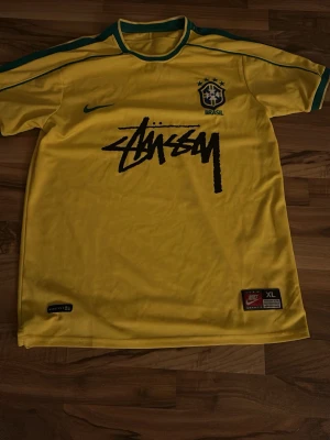 Gul Brasil x Stüssy t-shirt Nike - Gul Brasil t-shirt i samarbete mellan Nike och Stüssy. Klassisk fotbollströja med gröna detaljer, broderad CBF-logga och Stüssy-tryck på bröstet. Tillverkad i lätt och luftig polyester, perfekt för dig som gillar streetwear och sportig stil.