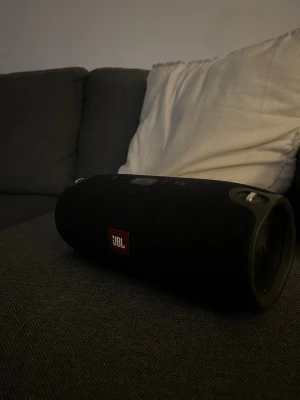 JBL svart bärbar högtalare - Kraftfull bärbar högtalare från JBL i svart med cylinderformad design och robust tygmaterial. Högtalaren har tydliga knappar på ovansidan och metallhandtag på sidorna för enkel transport. Perfekt för att ta med sig musiken överallt.