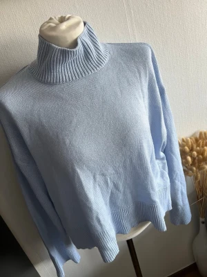 Ljusblå polotröja stickad - Säljer en ljusblå stickad polotröja med ribbad hög krage och muddar. Tröjan har en avslappnad passform och långa ärmar, perfekt för kyliga dagar. Materialet känns mjukt och bekvämt mot huden.