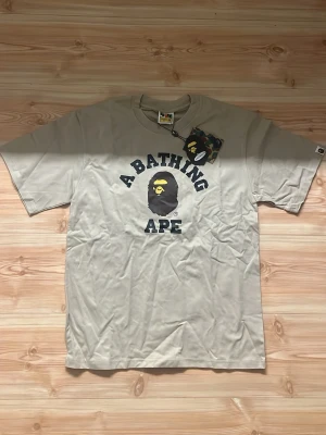 Bape t-shirt - Säljer en beige t-shirt från A Bathing Ape med det klassiska aptrycket och texten 'A BATHING APE' i svart på bröstet. T-shirten har rund hals, korta ärmar och är gjord i mjuk bomull. Perfekt för dig som gillar streetwear och vill sticka ut.