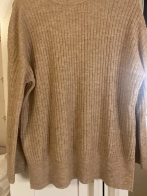 Beige ribbstickad oversized tröja - Mysig beige ribbstickad tröja med rund hals och långa ärmar. Tröjan har en avslappnad, oversized passform och slitsar i sidorna för extra komfort. Perfekt för lager på lager och enkel att matcha med olika outfits.