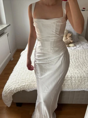 Vit långklänning i satin från BABYBOO med korsett  - Elegant vit långklänning i glansigt satinmaterial med breda axelband och figurnära passform. Klänningen har en stilren, enkel design med draperad överdel och markerad midja. Perfekt för speciella tillfällen som bal / bröllop. 