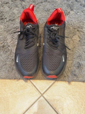 Nike Air Max 270  - Nike Air Max 270 sneakers i svart mesh med röda detaljer och vit logga. Skorna har en synlig Air-enhet i hälen, lågt skaft och snörning. Perfekta för dig som gillar sportig stil och vill ha bekväma skor med cool design.