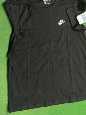 Svart Nike t-shirt med logga - Klassisk svart t-shirt från Nike med vit logga på bröstet. T-shirten har rund halsringning och korta ärmar. Perfekt basplagg för en sportig och avslappnad stil.