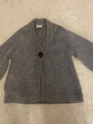 Grå kofta med knapp från Boomerang - Mysig grå kofta från Boomerang med en stor dekorativ knapp framtill. Koftan har bred ribbstickad krage och muddar, samt en avslappnad passform. Perfekt för lager på lager och enkel att matcha med olika outfits.