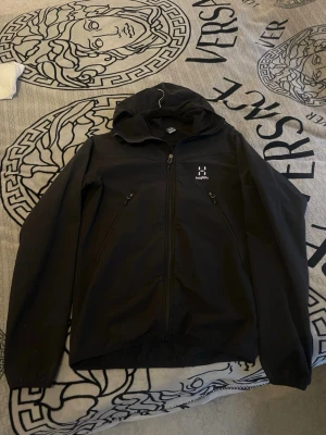 Svart Haglöfs vindjacka Apex Hood S - Svart vindjacka från Haglöfs, modell Apex Hood Men. Jackan har huva, två sidofickor med dragkedja och ventilerande meshfoder på insidan. Perfekt för blåsiga dagar och outdoor-aktiviteter. Snygg minimalistisk design med Haglöfs-logga på bröstet.