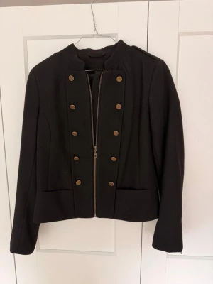 Svart vår jacka - Stilren svart jacka med militärinspirerade detaljer, dubbelknäppning med guldfärgade knappar och dragkedja framtill. Rak passform, axelklaffar och ståkrage ger en cool look. Långa ärmar med dragkedja vid ärmslut.